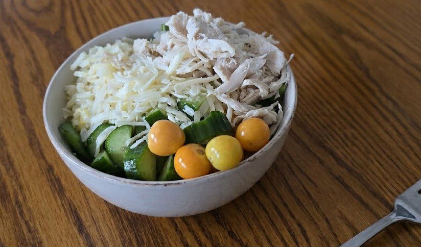 A high-angle close-up photograph of a chicken and cheese power bowl served on an oak wood table. The bowl, in a light grey ceramic, is piled with shredded poached chicken, shredded white cheese, diced cucumbers, and a small cluster of ripe golden gooseberries.