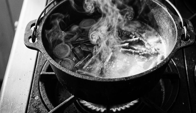 High-contrast black-and-white top-down photo of a cast-iron pot simmering on a gas stove. Wisps of steam rise from a broth containing sliced leeks and asparagus, emphasizing the analog ritual of preparing gut-health comfort foods.