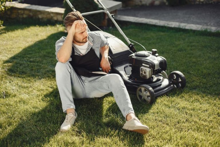 tired homeowner in a black apron sitting on grass, looking exhausted after using a manual lawn mower. Low maintenance landscaping can prevent this exhaustion.