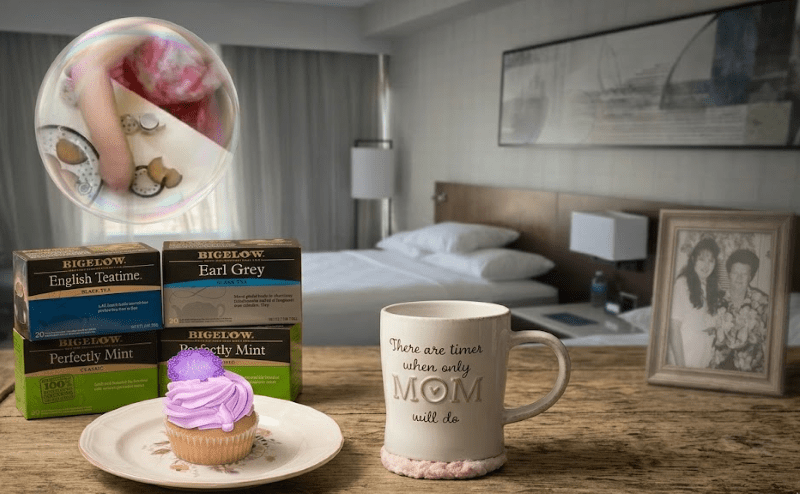 A close-up photograph on a rustic wooden table featuring a purple frosted cupcake on a plate next to a white mug that reads "There are times when only MOM will do." Behind them are stacks of Bigelow tea boxes (English Teatime, Earl Grey, Perfectly Mint), a small gold spoon, and loose tea bags. The out-of-focus background shows a hotel room with two beds and a framed black and white photo of a woman and child. A large, floating bubble in the foreground contains a small hand and tiny ceramic dishes.